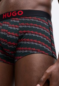 Boxeri negri din material moale cu un model repetitiv de logo "Hugo" în roșu și gri. Bretelele sunt negre, cu litere îndrăznețe în roșu.