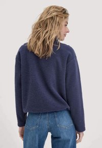 Frau mit blonden, welligen Haaren, die einen marineblauen Pullover und blaue Jeans trägt, steht mit dem Rücken zu einem schlichten, hellen Hintergrund.