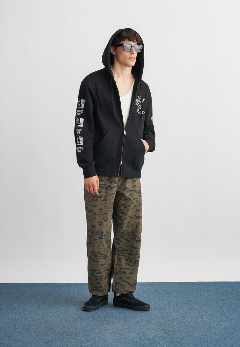 Junger Mann trägt schwarzen Kapuzenpullover mit Reißverschluss und weißen Grafiken, Camouflage-Muster Hose, schwarze Turnschuhe und dunkle Sonnenbrille, steht auf blauem Boden.