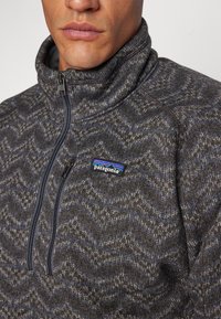 Patagonia fleece pullover, gray with blue zigzag pattern, high collar, quarter-zip front, and logo on left chest. Soft texture.