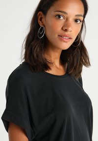 Black t-shirt with short rolled sleeves, featuring a relaxed fit and smooth texture. Model wears silver hoop earrings for accent.
