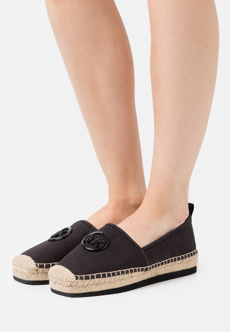 Jambes portant des espadrilles en toile noire avec semelle en jute tressée et logo rond noir sur le dessus, sur fond blanc.
