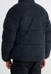 Tmavě šedá fleecová prošívaná bunda s texturovaným, měkkým povrchem. Má vysoký límec a vodorovné prošívané panely. Zobrazený pohled zezadu.