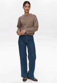 Maglione a maglia marrone con maniche lunghe, abbinato a jeans larghi a strisce verticali blu. La modella indossa scarpe nere in tessuto piatte.