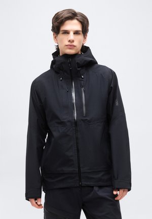 Νεαρός άνδρας που φοράει μαύρο αδιάβροχο μπουφάν με κουκούλα και φερμουάρ μπροστά, στέκεται μπροστά σε απλό ανοιχτό φόντο.