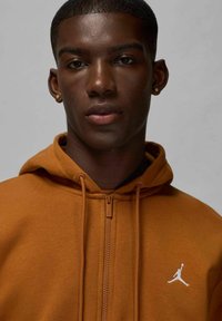 Felpa con zip marrone con finitura testurizzata. Presenta un cappuccio con cordino e un logo Jumpman bianco sul lato sinistro del petto. Tessuto liscio.