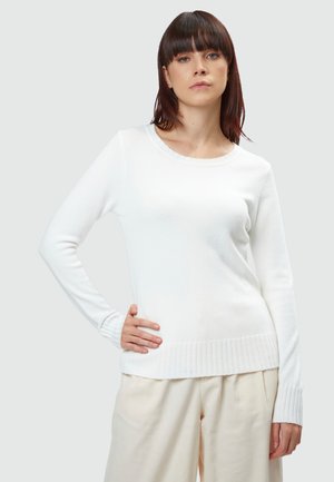 Maglione bianco a maniche lunghe in materiale morbido con colletto rotondo, polsini e orlo a coste. Indossato con pantaloni chiari e larghi.
