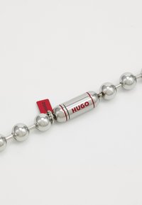 Metallperle armring med blanke sølvkuleperler og en sentral kapselcharms merket "HUGO" med røde og sølvfargede detaljer.
