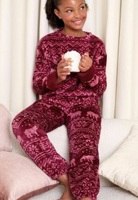 Conjunto de pijama de polar burdeos con un patrón de fair isle y osos polares. Textura suave, puños acanalados y corte relajado. Sostiene una taza blanca con un diseño de galleta de jengibre.