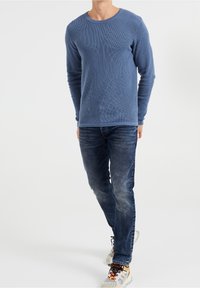 Blå långärmad crewneck-tröja med ett texturerat mönster, bärs tillsammans med mörka jeans och flerfärgade sneakers.