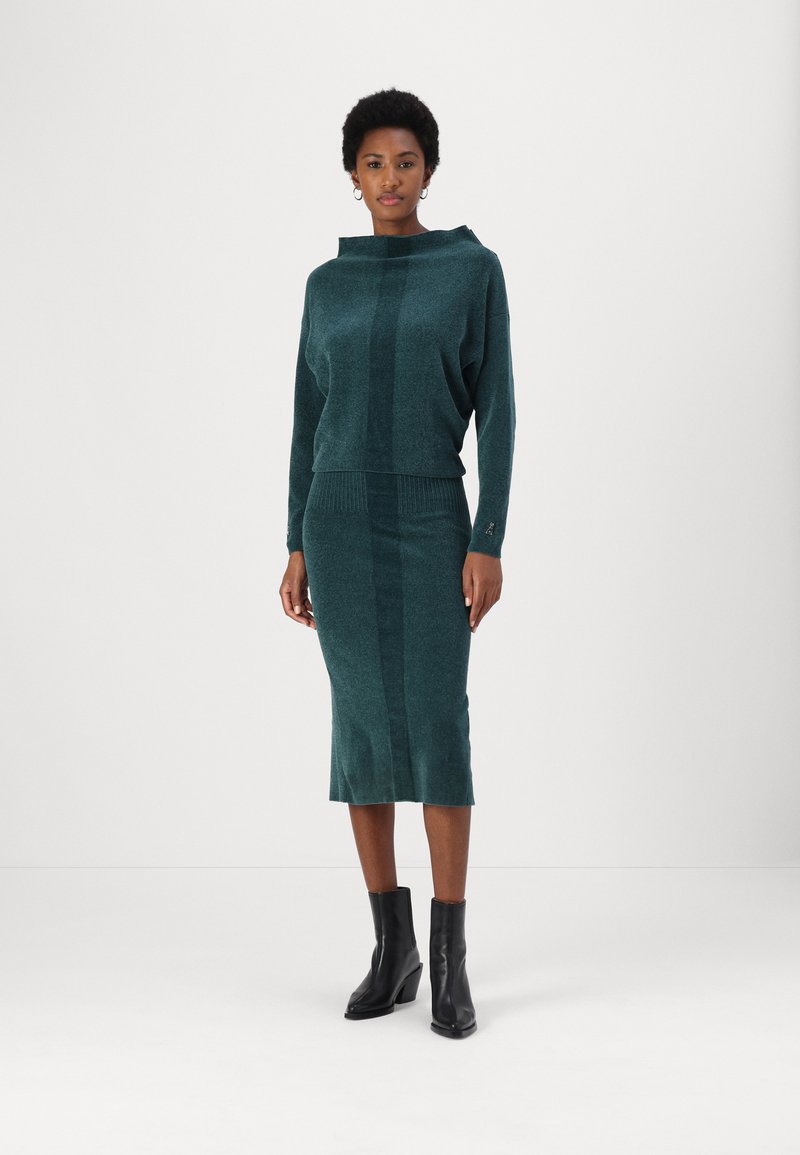 Teal gebreide trui met een brede halslijn, gecombineerd met een bijpassende kokerrok. Beide hebben een subtiele textuur en verticale strepen. Zwarte enkelboots.