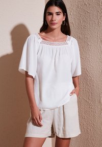 White blouse with puff sleeves and lace detailing at the neckline, paired with light beige linen shorts. Smooth texture, loose fit.