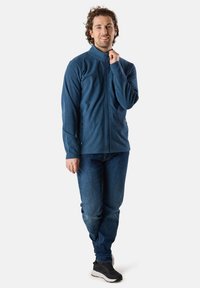 Mörkblå vindjacka med dragkedja och hög krage, dubbla bröstfickor, i kombination med denimjeans och svarta sneakers. Slät textur, avslappnad passform.