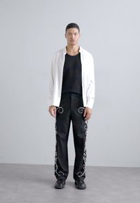 Fekete nadrág fehér virágos hímzéssel az oldalán, fekete ujjatlan felsővel és egy nyitott fehér inggel párosítva. Sima anyag és laza fazon.