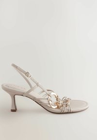 Beige Absatzsandale mit Riemchen-Design, einem goldenen Kettenakzent an der Vorderseite und einem schlanken Absatz. Geschmeidige Textur mit geschlossener Zehenpartie.