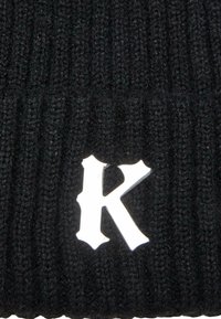 Fekete kötött sapka függőleges bordázott textúrával és fehér, stilizált "K" díszítéssel az elején.
