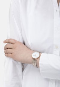 Runde Uhr mit einem weißen Zifferblatt, goldfarbenen Akzenten und einem silbernen Metallarmband. Wird an einer Hand getragen, die auf einem weißen Hemdsärmel ruht.