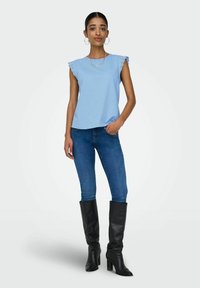 Helles blaues ärmelloses Oberteil mit Spitzendetails, kombiniert mit dunklen Denim-Jeans und knielangen schwarzen Lederstiefeln, vor einem neutralen Hintergrund.