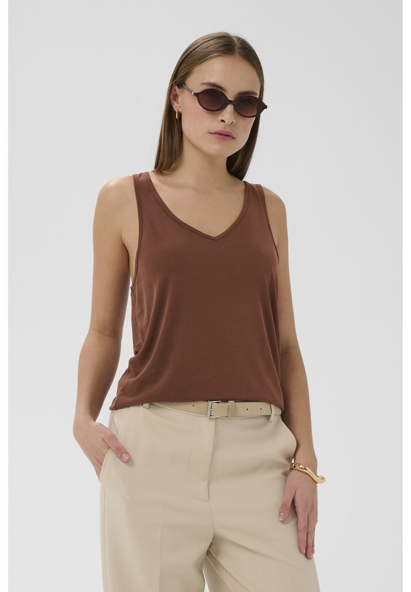 Mujer vestida con top marrón sin mangas, pantalones beige con cinturón, gafas de sol, pulsera de oro y pendientes, de pie con una mano en el bolsillo.