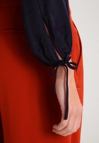 Haut à manches longues bleu marine avec un poignet élastique présentant une fente et un détail noué. Un tissu rouge texturé est visible en arrière-plan.