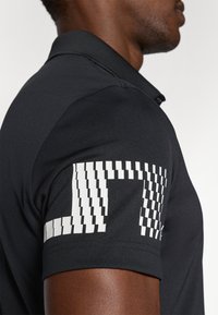 Polo preto com colarinho estruturado e um distinto padrão xadrez branco na manga esquerda. Tecido liso e elástico.