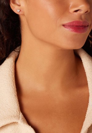 Boucle d'oreille en argent en forme de coeur, portée sur l'oreille d'un modèle ; une peau lisse, un maquillage léger et un haut crémeux texturé sont visibles.