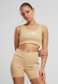 Beige idrettslig crop topp og høyhælte shorts laget av glatt stoff med "KANI"-logo i hvitt. Fitted design med en minimalistisk stil.