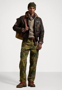 Brązowa skórzana kurtka bomber z futrzanym kołnierzem, oliwkowo-zielona dzianinowa czapka, szary sweter, koszula w kratę, spodnie moro i brązowe buty. Brązowa torba na ramię.