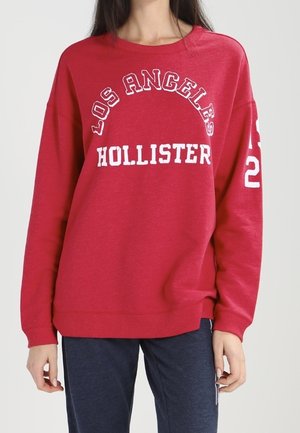 Rode sweatshirt met "LOS ANGELES" en "HOLLISTER" in witte tekst, geribbelde manchetten en onderrand, ontspannen pasvorm, zachte stof, ronde hals.