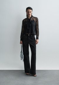 Femme portant un blouse noir transparent à manches longues avec nœud, un pantalon large noir, des sandales noires, tenant un sac à main argenté avec des pampilles.