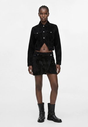 Femme debout avec des cheveux tressés portant une veste courte noire, une jupe portefeuille noire et des bottines noires sur fond blanc.