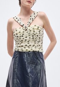 Top crem cu buline și detalii strânse, cu bretele late legate; asortat cu o fustă închisă la culoare, lucioasă și cu textură.