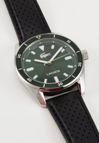 Relógio Lacoste com um mostrador verde, caixa em aço inoxidável prateada, marcadores de horas brancos, bracelete de couro perfurado preto e uma coroa de lado.