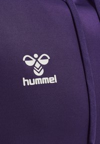 Top sportivo viola con finitura testurizzata, dotato di un logo "hummel" bianco e un design distintivo di ape sopra il nome del marchio.
