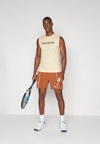 Cremefarvet ærmeløs top med sort tekst, brune shorts med hvide detaljer, holder en tennisracket og bold, iført hvide sneakers.