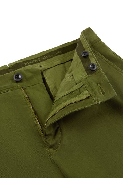 Olijfgroene broek met gedeeltelijk open ritssluiting, twee zwarte knopen op de tailleband en zichtbare knoopsgaten.
