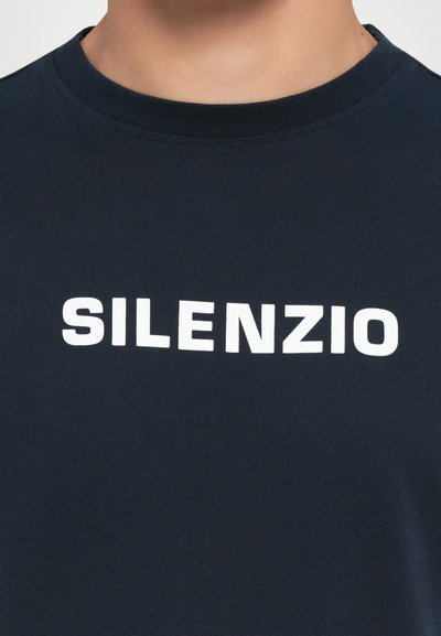 Tumesinise lühikese varrukaga T-särk, ümmarguse kaeluse ja ette trükitud sõnaga "SILENZIO" rasvases, valges suurkirjas.