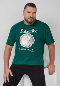 Mężczyzna z brodą noszący zieloną koszulkę z białym graficznym wzorem pierścienia drzewa i napisem "Subskrybuj Naturę" siedzi z ręką opartą na białym bloku.