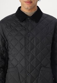 Casaco acolchoado preto com colarinho de veludo, botões de pressão, um bolso com marcação em relevo e textura com padrão em diamante.