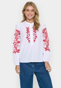 Hvid bluse med rød blomsterbroderi på ærmerne, knaplukning og elastiske manchetter. Kombination med blå denimbukser.