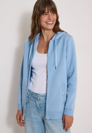 Femme souriante portant un sweat à capuche bleu clair zippé sur un débardeur blanc et un jean bleu clair, debout devant un fond uni.