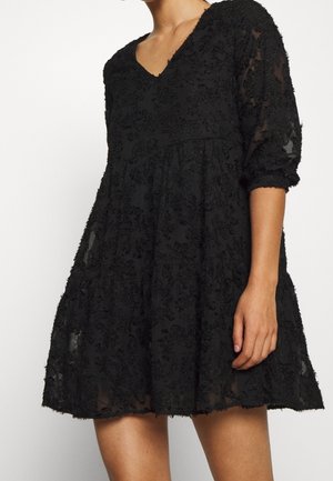 Robe noire avec décolleté en V, manches trois-quarts, et tissu à texture florale. Silhouette évasée avec design en couches et accents transparents.