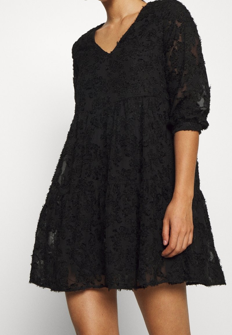 Robe noire avec décolleté en V, manches trois-quarts, et tissu à texture florale. Silhouette évasée avec design en couches et accents transparents.