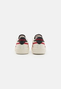 White sneakers with red and black accents, featuring a textured sole, smooth leather upper, and "Reebok" branding at the heel.