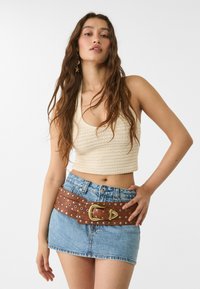 Béžový pletený cropped top, modrá denimová mini sukně a široký hnědý opasek s kovovými cvočky a zlatou sponou, se stříbrnými a zlatými cvočky.