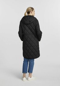 Czarna pikowana długa kurtka z kapturem, zwężona w talii, o miękkiej fakturze. Nosi ją z niebieskimi dżinsami i białymi trampkami.