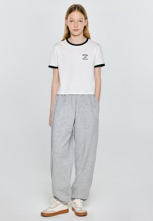 Młoda osoba stojąca, ubrana w biały krótki top z czarną lamówką, luźne szare spodnie dresowe i beżowe tenisówki na jednolitym tle.