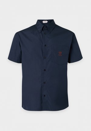 Jeune homme portant une chemise bleu marine à manches courtes boutonnée, un jean bleu délavé et des chaussures noires avec des accents dorés, debout devant un fond uni.
