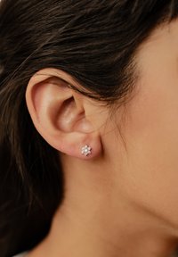 Pendiente de plata con forma de flor, adornado con múltiples pequeños cristales transparentes, situado en primer plano junto a la oreja de una persona y con el cabello oscuro de fondo.