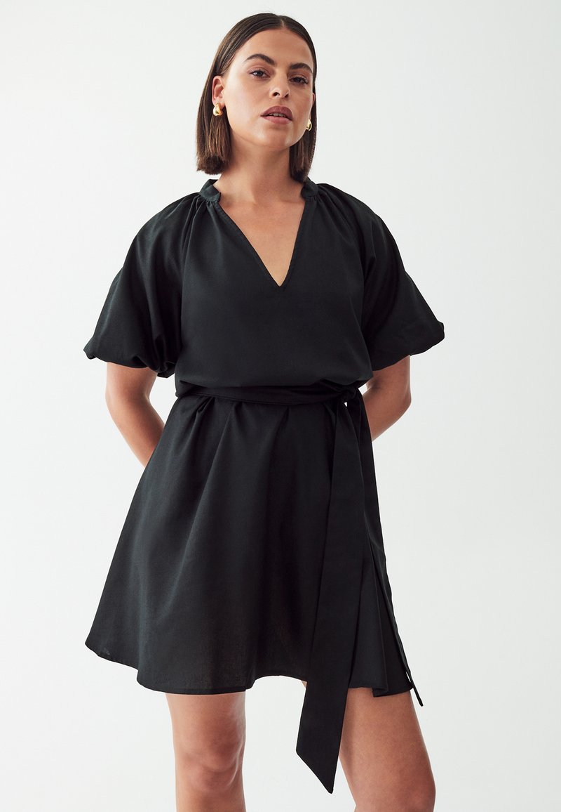 Zwarte jurk met een V-hals, korte poffende mouwen, een cinched taille en een strikceintuur. De stof heeft een gladde textuur en een vloeiende uitstraling.